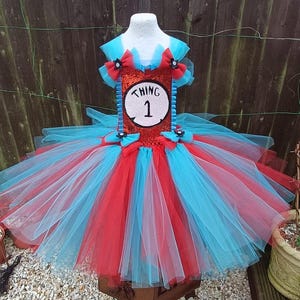 May include: A red, white, and blue tutu dress with the text "THING 1" on the bodice. The dress features a red sequined top, blue tulle sleeves, and a layered skirt of red and blue tulle. Black bows accent the dress.