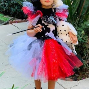 May include: A child's Cruella de Vil costume, with a black and white wig, sunglasses, and a tutu. The tutu is red, white, and black with tulle layers. The child holds a wand and a stuffed Dalmatian dog.