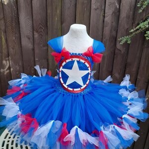May include: A blue, red, and white tutu dress with a Captain America shield design. The bodice features a shield with a white star on a blue background, surrounded by a red border. The skirt is layered with blue, red, and white tulle.
