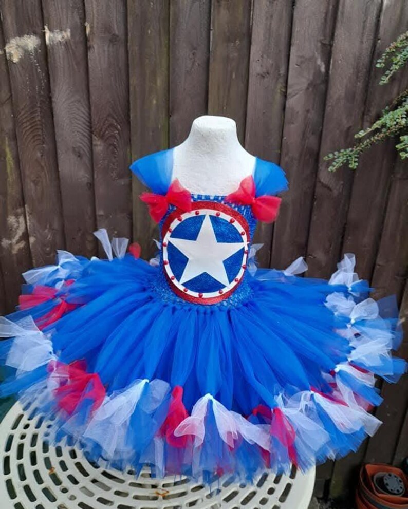 May include: A blue, red, and white tutu dress with a Captain America shield design. The bodice features a shield with a white star on a blue background, surrounded by a red border. The skirt is layered with blue, red, and white tulle.