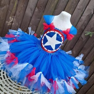 May include: A blue, red, and white tutu dress with a star emblem. The bodice features a round emblem with a white star on a blue background, trimmed in red. The dress has a layered tulle skirt with red and white accents, and blue shoulder straps with red bows.