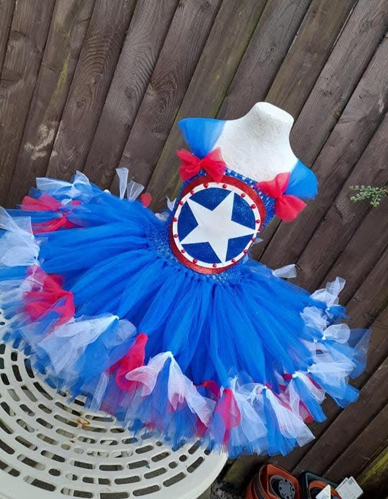 May include: A blue, red, and white tutu dress with a star emblem. The bodice features a round emblem with a white star on a blue background, trimmed in red. The dress has a layered tulle skirt with red and white accents, and blue shoulder straps with red bows.