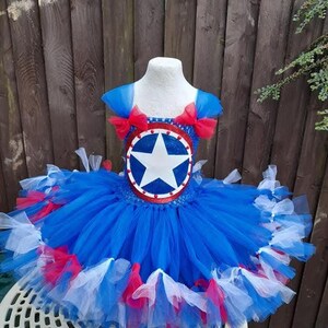 May include: A blue, red, and white tutu dress with a star emblem on the bodice. The dress features a layered tulle skirt and red bows on the shoulders. The bodice has a circular emblem with a white star.
