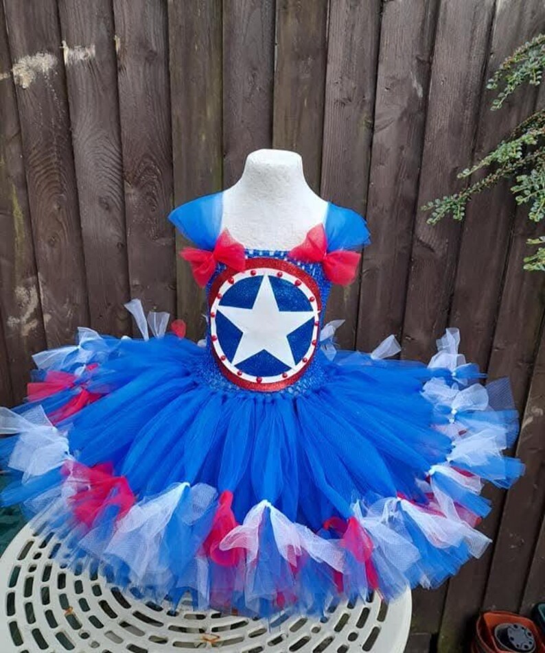 May include: A blue, red, and white tutu dress with a star emblem on the bodice. The dress features a layered tulle skirt and red bows on the shoulders. The bodice has a circular emblem with a white star.