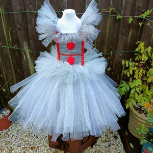 May include: A clown-themed tutu dress in white and grey, displayed on a mannequin. The bodice is grey with red detailing and large red circles. The skirt is made of layers of white tulle with glitter, creating a fluffy effect.