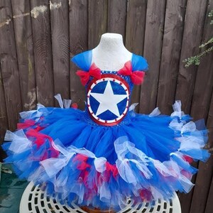 May include: A blue, red, and white tutu dress with a star emblem. The dress features a blue bodice with red bows on the shoulders and a layered skirt with red and white accents. The dress is displayed on a mannequin.