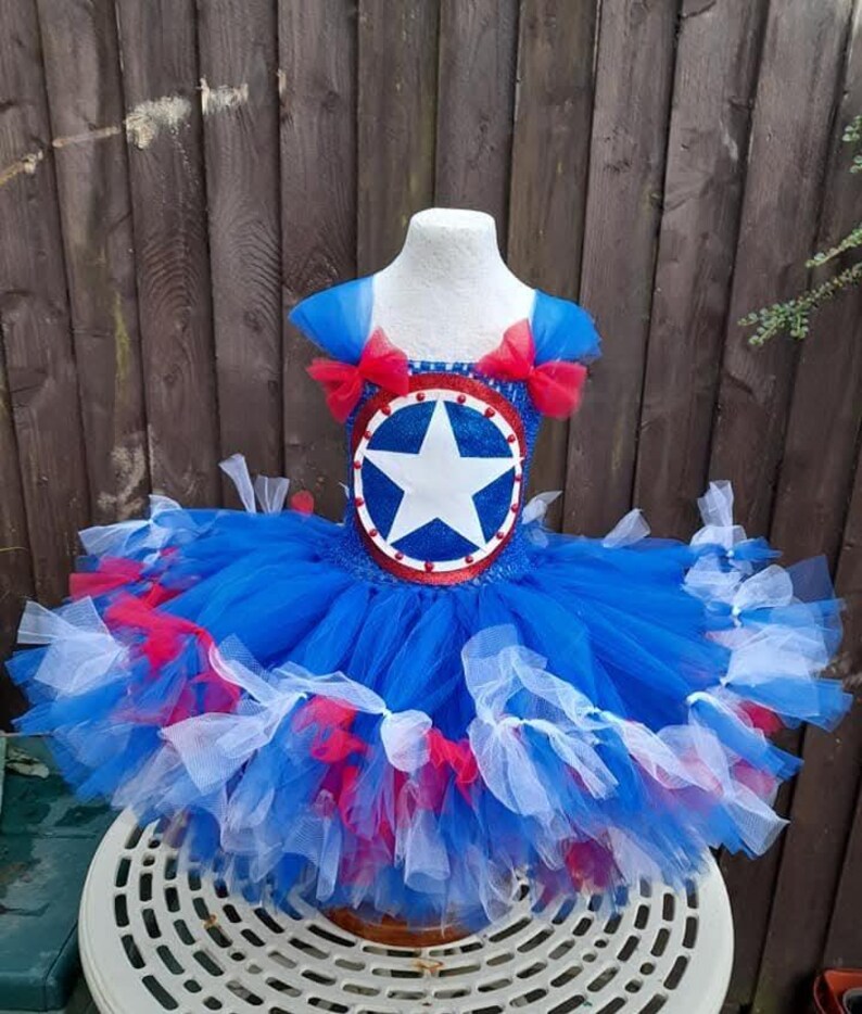 May include: A blue, red, and white tutu dress with a star emblem. The dress features a blue bodice with red bows on the shoulders and a layered skirt with red and white accents. The dress is displayed on a mannequin.