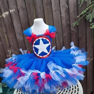 May include: A blue, red, and white tutu dress with a star emblem on the bodice. The dress features a layered tulle skirt and red bows on the shoulders. The bodice has a circular emblem with a white star.