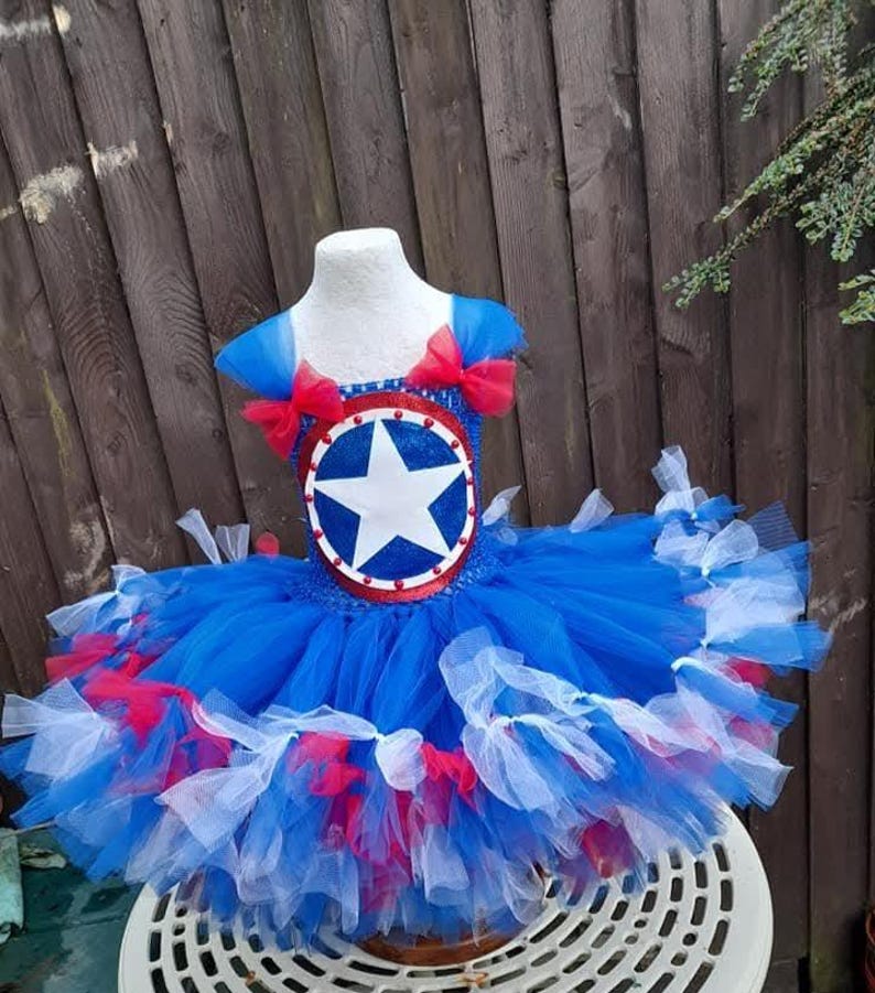May include: A blue, red, and white tutu dress with a star emblem on the bodice. The dress features a layered tulle skirt and red bows on the shoulders. The bodice has a circular emblem with a white star.