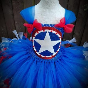 May include: A blue, red, and white tutu dress with a star emblem. The bodice features a circular emblem with a white star on a blue background, trimmed with red. The tutu skirt is layered with blue, red, and white tulle, and the shoulders are adorned with red bows.