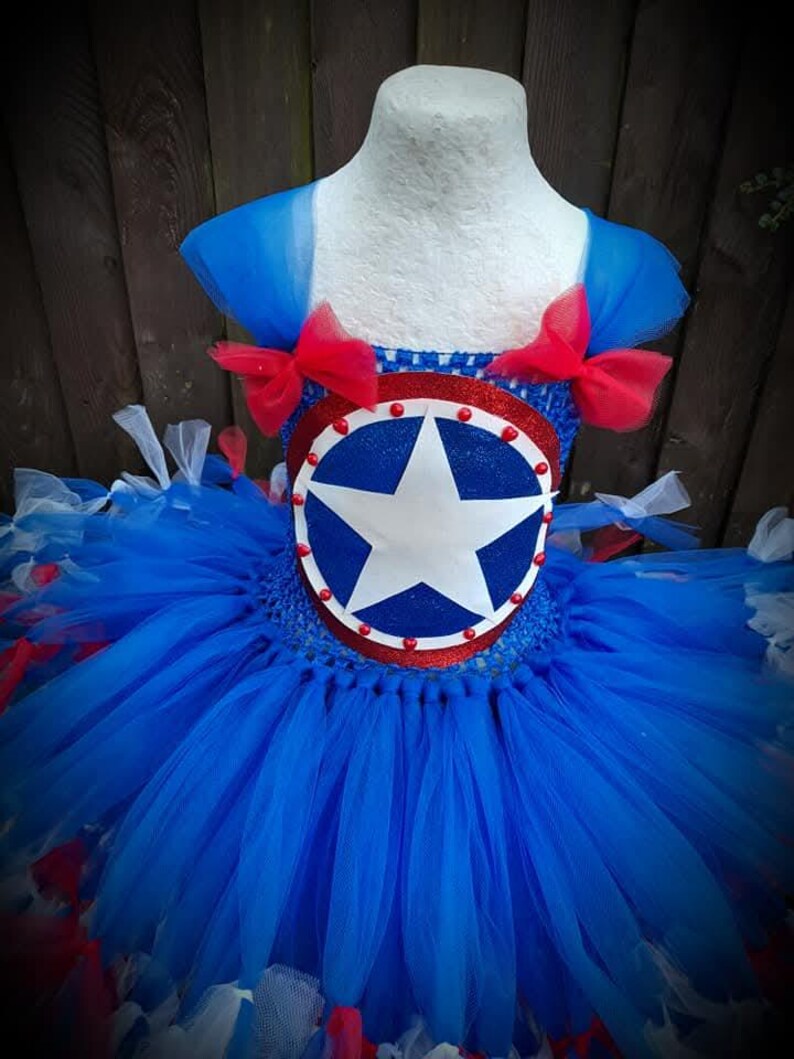May include: A blue, red, and white tutu dress with a star emblem. The bodice features a circular emblem with a white star on a blue background, trimmed with red. The tutu skirt is layered with blue, red, and white tulle, and the shoulders are adorned with red bows.