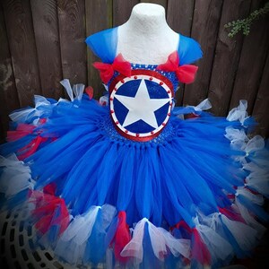 May include: A blue, red, and white tutu dress with a Captain America shield design on the bodice. The dress features layers of blue tulle, red bows, and accents of red and white tulle. The bodice has a blue and red shield with a white star.