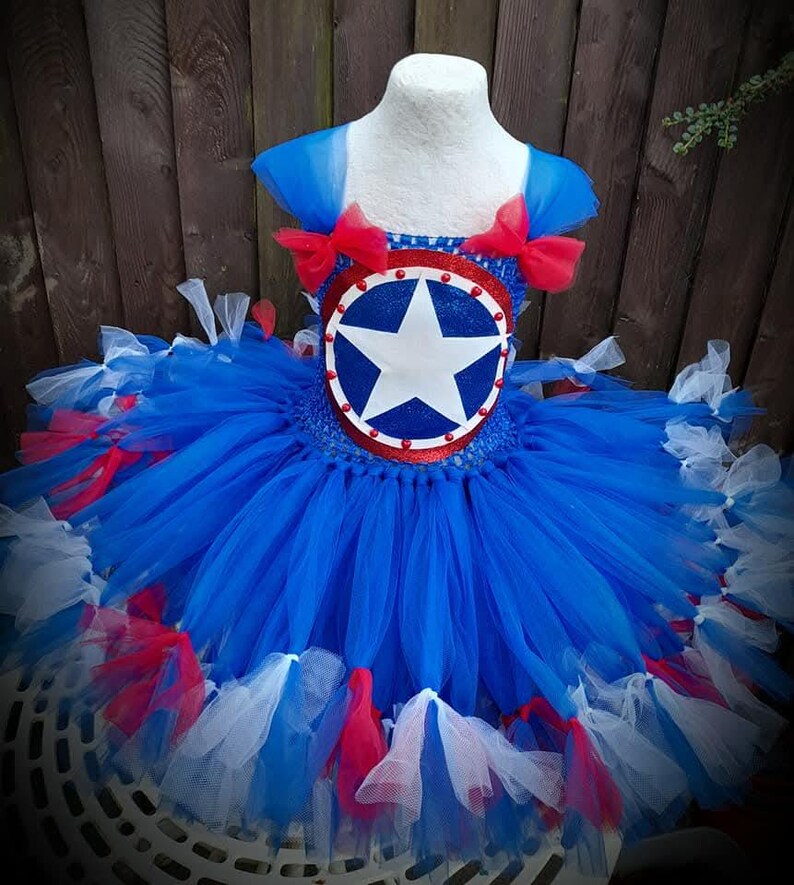 May include: A blue, red, and white tutu dress with a Captain America shield design on the bodice. The dress features layers of blue tulle, red bows, and accents of red and white tulle. The bodice has a blue and red shield with a white star.