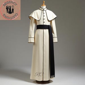 Może przedstawiać: Kremowa suknia wizytowa z czarną szarfą i wykończeniami. Suknia posiada pelerynę, wysoki kołnierz i czarne guziki z przodu. Długie rękawy mają czarne wykończenia na mankietach. Suknia jest prezentowana na manekinie.