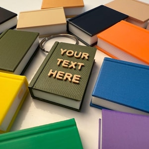 May include: A collection of small book-shaped keyrings in various colours, including green, blue, yellow, orange, and purple. One keyring has the text "YOUR TEXT HERE" on the cover.
