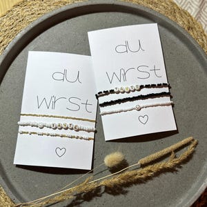 Könnte beinhalten: Zwei weiße Karten mit schwarzem Text, der "du wirst" lautet, und einem Herzsymbol. Jede Karte hat ein Perlenarmband mit dem Wort "Tante" in goldenen Perlen. Das Armband auf der oberen Karte ist schwarz-weiß, und das Armband auf der unteren Karte ist gold-weiß.