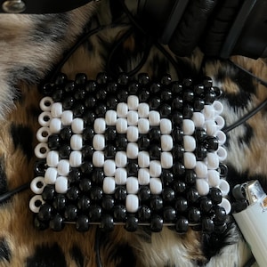 Può includere: Un portachiavi a forma quadrata con perline nere e bianche e un motivo geometrico. Il portachiavi è realizzato con piccole perline rotonde che sono infilate insieme per creare una forma quadrata.