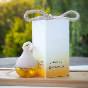 May include: A wooden and glass essential oil diffuser with a yellow liquid inside, next to a white box with the words "Jungle Escapade" printed on it. The box has a gold and yellow gradient. A rope is tied to the top of the box.