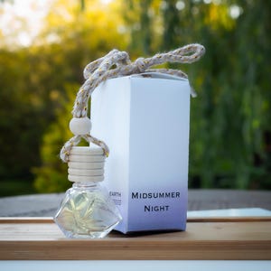May include: A clear, diamond-shaped glass bottle with a wooden top and a decorative rope. The bottle hangs in front of a white box with the words "Midsummer Night" printed on it. The background is blurred with green and yellow tones.