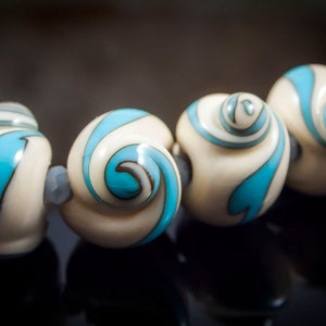 May include: Close-up of a strand of cream-colored beads with a spiral design in turquoise and white. The beads are strung together with small gray beads, creating a unique jewelry piece.