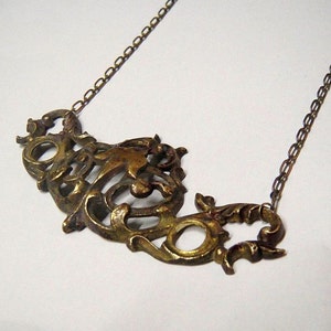 May include: A gold-toned necklace with a large, ornate pendant featuring a stylized design. The pendant is made of metal and has a detailed, intricate pattern.