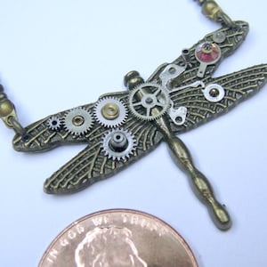 May include: A steampunk-style dragonfly pendant necklace. The bronze-toned metal dragonfly features intricate gear details and a small red gemstone. The necklace has a delicate chain.