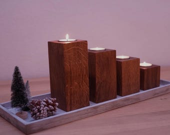 May include: Four wooden candle holders in a graduated size, arranged in a row on a gray tray. The holders are made of dark wood with a natural grain pattern. The candles are lit and casting a warm glow. There are two small artificial pine trees and a pine cone on the tray.