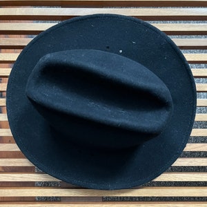 Peut inclure: Un chapeau fedora en feutre noir avec un large bord et une calotte légèrement enfoncée.