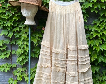 May include: A white cotton tiered skirt with a gathered waistband. The skirt is hanging on a wooden hanger against a backdrop of green ivy.