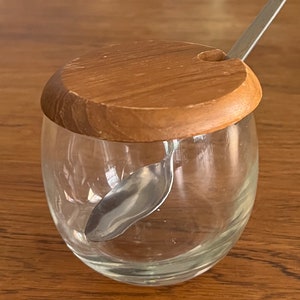 May include: A clear glass jar with a wooden lid and a small metal spoon inside. The lid has a hole for the spoon to fit through.