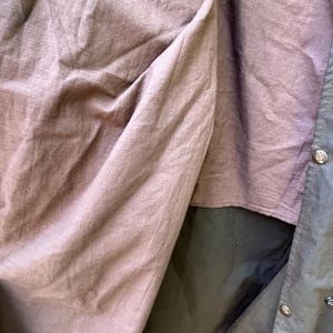 May include: Close-up of a dark olive green jacket interior with a mauve-colored lining. The jacket has a button closure and a dark green exterior. The lining is made of a soft, textured fabric.
