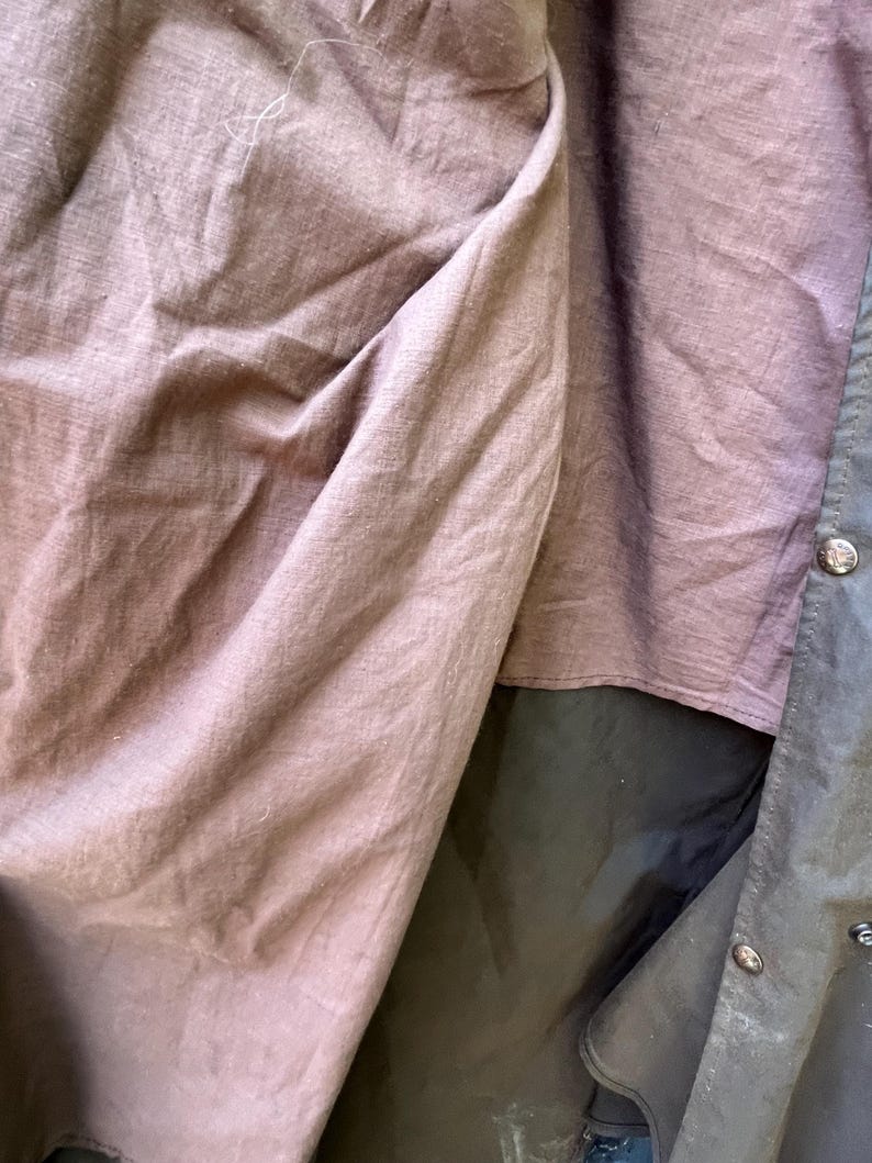 May include: Close-up of a dark olive green jacket interior with a mauve-colored lining. The jacket has a button closure and a dark green exterior. The lining is made of a soft, textured fabric.