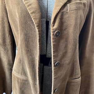 Op de afbeelding: Een bruine corduroy blazer met een revers, drie knopen en twee voorzakken. De jas wordt op een mannequin getoond. De corduroy stof heeft een verticale ribstructuur.