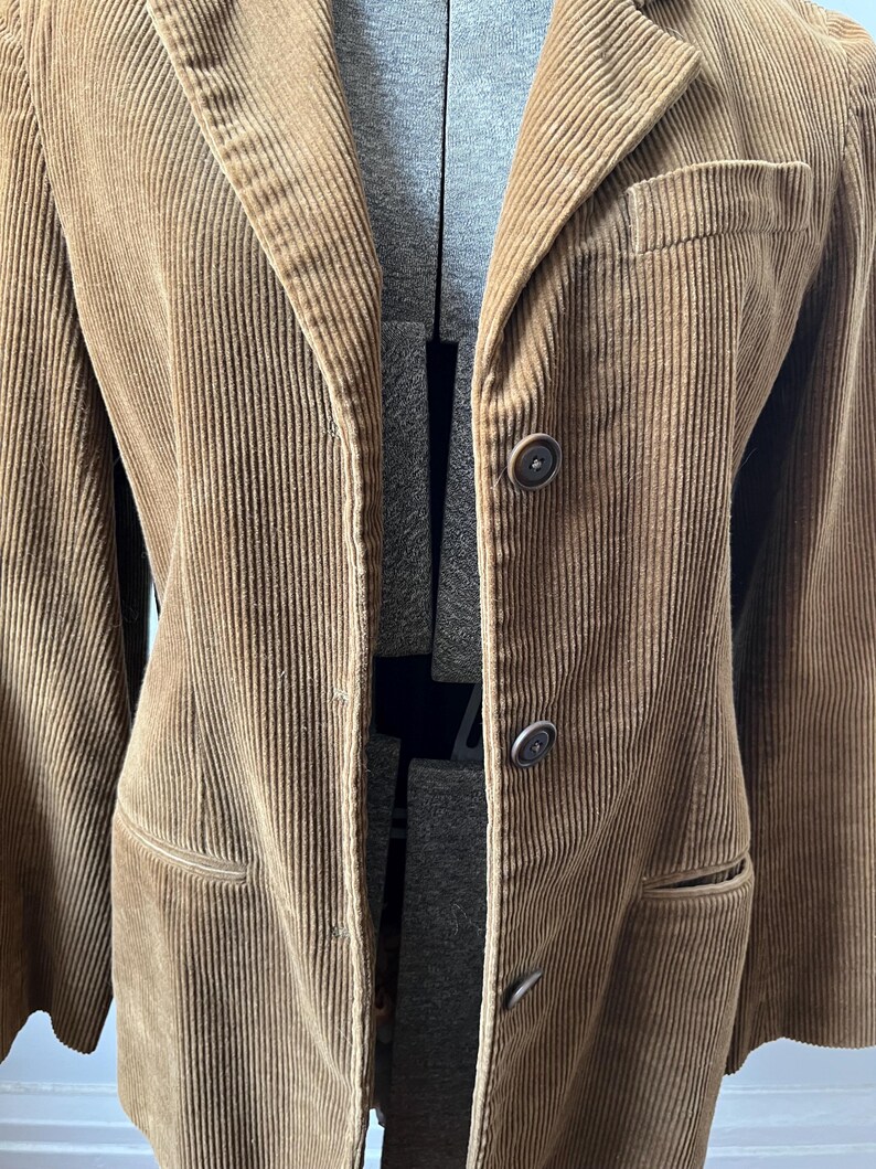 Op de afbeelding: Een bruine corduroy blazer met een revers, drie knopen en twee voorzakken. De jas wordt op een mannequin getoond. De corduroy stof heeft een verticale ribstructuur.