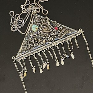 May include: Silver-toned triangular pendant necklace with intricate floral and scrollwork designs. Features a turquoise stone and a small red gem. The pendant has dangling chains with small, diamond-shaped charms. The necklace chain is a delicate box chain.