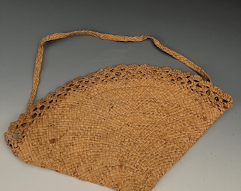 May include: A tan-coloured woven straw bag with a semi-circular shape and a braided handle. The bag has an open-weave border along the top edge. The bag is made of natural fibres.