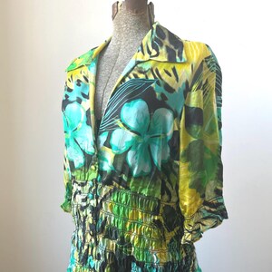 May include: A colorful floral print blouse with a collared neckline and three-quarter sleeves. The blouse features a vibrant pattern of green, yellow, and turquoise flowers and leaves. The waistline is gathered with elastic.