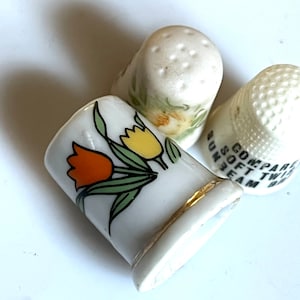 Op de afbeelding: Drie witte porseleinen vingerhoedjes. Een vingerhoedje heeft een bloemenpatroon met gele en oranje tulpen. Een ander vingerhoedje heeft een bloemenpatroon met gele bloemen. Het derde vingerhoedje heeft zwarte tekst met de tekst "COMPARE SOFT TWIN SUNBEAM".