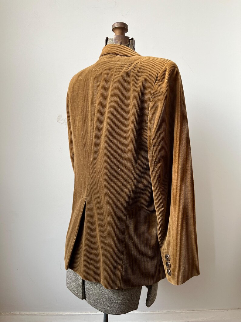 Op de afbeelding: Bruine corduroy blazer met een gekerfde revers en drie knopen op elke manchet. De blazer wordt getoond op een mannequin, met de achterkant en het zijprofiel. De stof heeft een geribbelde textuur.