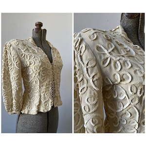 May include: A cream-colored jacket with a V-neck and long sleeves. The jacket is covered in intricate white embroidery that resembles floral patterns.