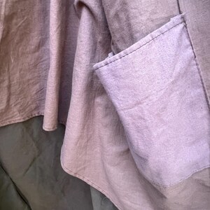 May include: Close-up of a garment featuring a muted purple fabric with a visible pocket. The fabric has a textured appearance, with visible seams and a layered design. The garment appears to be made of a soft, flowing material.