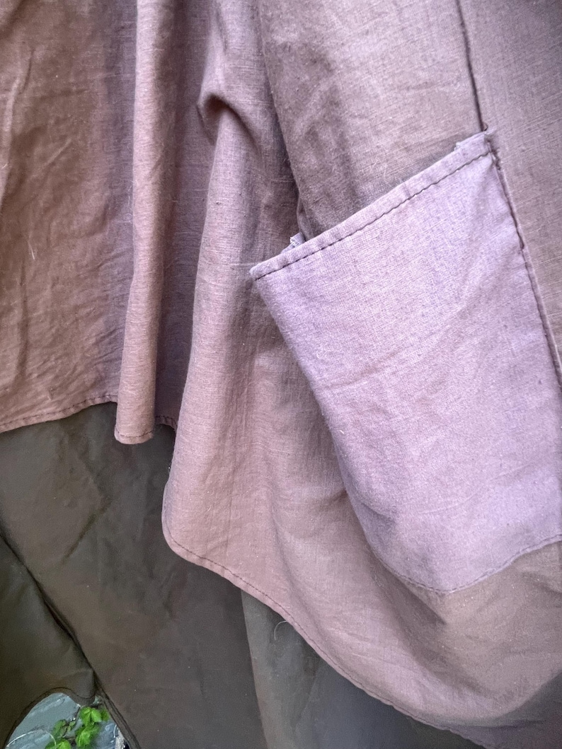 May include: Close-up of a garment featuring a muted purple fabric with a visible pocket. The fabric has a textured appearance, with visible seams and a layered design. The garment appears to be made of a soft, flowing material.