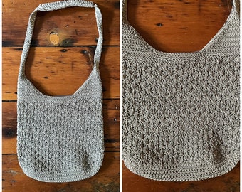 Könnte beinhalten: Eine hellgraue gehäkelte Tragetasche mit einem langen Riemen. Die Tasche hat ein schlichtes, offenes Design und ist aus einem leichten Material gefertigt.