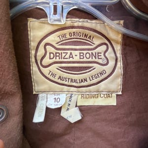 May include: Close-up of a brown riding coat label. The label reads "THE ORIGINAL DRIZA-BONE THE AUSTRALIAN LEGEND." The size is 10, with a chest measurement of 130 cm. The coat is made of 100% cotton.