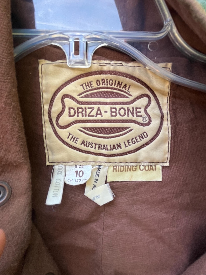 May include: Close-up of a brown riding coat label. The label reads "THE ORIGINAL DRIZA-BONE THE AUSTRALIAN LEGEND." The size is 10, with a chest measurement of 130 cm. The coat is made of 100% cotton.