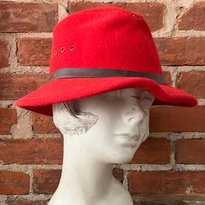 Puede incluir: Un sombrero fedora de fieltro rojo con una banda de cuero marr&oacute;n y una ala que se curva hacia abajo. El sombrero tiene tres peque&ntilde;os agujeros en la corona.