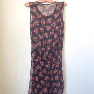 May include: A sleeveless, sheer dress with a dark background and a floral pattern in shades of pink and red. The dress hangs on a wooden hanger, showcasing its flowing, lightweight fabric and asymmetrical hemline.