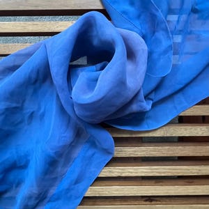 May include: A vibrant blue scarf made of sheer fabric, draped over a wooden slatted surface. The scarf is folded and gathered, showcasing its flowing texture and color. The edges are finished with a delicate stitch.