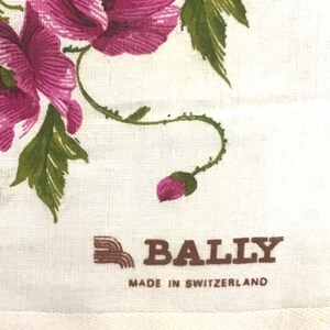 Pu&ograve; includere: Un tessuto bianco con un motivo floreale rosa e un logo Bally marrone con il testo "Made in Switzerland".