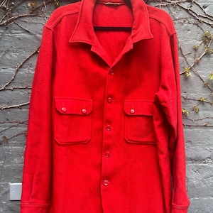 May include: A bright red button-down shirt with two chest pockets. The shirt has a collar and long sleeves. The garment is hanging on a black hanger against a gray brick wall with vines.